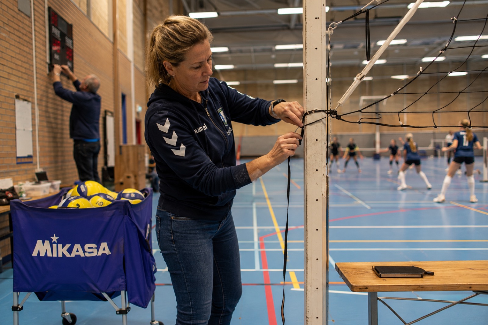 Iemand spant een volleybalnet terwijl op de achtergrond gespeeld wordt